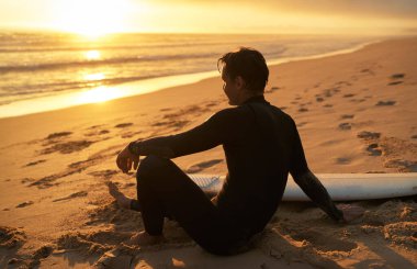 Bir sörfçü gün batımında, sörf tahtasının yanında, huzurlu okyanus manzarasının tadını çıkarırken, plajda biraz dinlenir..
