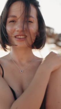 Kısa siyah saçlı ve siyah elbiseli güzel bir kadın, güneşli bir sahilde saçları rüzgarda uçuşurken gülümsüyor..