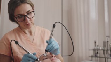 Odaklanmış estetikçi, modern bir güzellik kliniğinde özel ekipmanlar kullanarak gevşemiş bir müşteriye mikro akım yüz terapisi uygular..