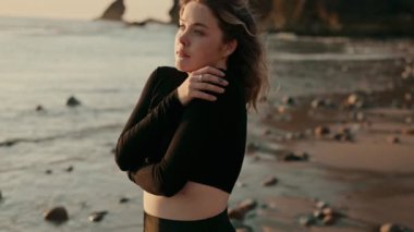 Genç bir kadın kayalık bir plajda duruyor, sakin bir gün batımına bakarken kendini kucaklıyor, sükuneti ve iç güzelliği yansıtıyor..