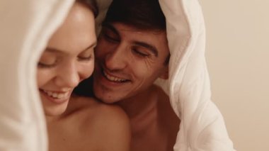 Romantik bir anı paylaşan güzel bir çift, yatakta rahat bir battaniyenin altında flört ediyor ve öpüşüyor, mutluluk ve aşk saçıyor..