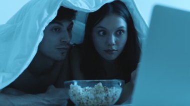 Genç çift evde film gecesinin tadını çıkarıyor, bir kase patlamış mısır ve dizüstü bilgisayarla battaniyenin altına kıvrılmış..