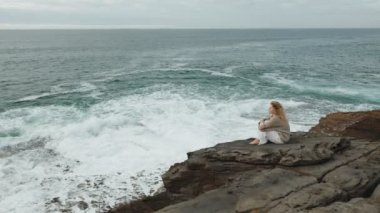 Düşünceli bir kadın kayalıklarda tek başına oturur ve sonsuz denize bakar. Yansımayı ve yalnızlığı sembolize eder..