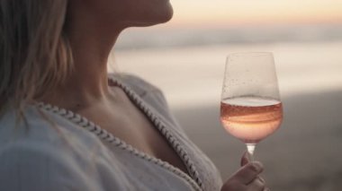 Beyaz elbiseli bir kadın gün batımında sahilde bir bardak gül şarabı içiyor. Bu video rahatlama, lüks ve yaz havasını çağrıştırıyor..