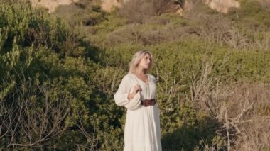 Beyaz elbiseli baştan çıkarıcı bir kadın doğanın güzelliğinin ortasında duruyor. Güneşi emerken gözleri kapalı..