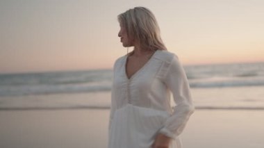 Beyaz bir elbise giyen, gün batımında kumlu bir plajda yürüyen uzun dalgalı saçlı güzel sarışın kadın. Okyanusun huzur ve sükunetinin tadını çıkarırken mutlu ve tasasız görünüyor..