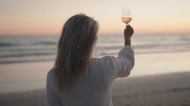 Sarışın bir kadın, kumsalda sıcak bir yaz akşamında bir bardak gül şarabıyla gün batımına kadeh kaldırıyor..