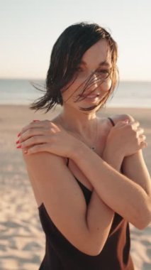 Siyah elbiseli güzel genç bir kadın gün batımında plajda rahatlatıcı bir günün tadını çıkarıyor. Yavaş çekim.