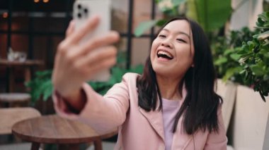 Pembe ceketli mutlu Asyalı kız açık hava kafeteryasında telefonuyla selfie çekiyor..