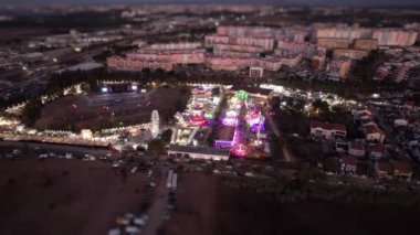 Geceleri kalabalık bir eğlence parkının havadan görüntüsü. Park renkli ışıklarla aydınlanıyor ve insanlar gezintilerin ve eğlencenin tadını çıkarıyorlar..