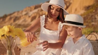 Bir anne güneşli bir plaj pikniğinde oğluna portakal suyu doldurur. İkisi de beyaz giyinmiş ve gülümsüyor. Masada sarı çiçekler var. Neşeli bir yaz havası katıyor..