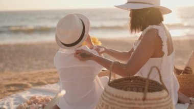 Bir anne, gün batımındaki plaj pikniğinde battaniyeye oturup, huzurlu ve mutlu bir atmosfer yaratan ılık güneşin ve hafif esintinin tadını çıkarırken oğluna meyve suyu verir.