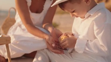 Bir anne ve küçük oğlu gün batımında plajda piknik yapıyorlar. İkisi de beyaz giyinmiş, mutlu ve rahat görünüyorlar..