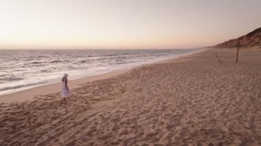 Gün batımında kumlu bir kumsalda yürüyen bir annenin, doğanın ve okyanusun güzelliğinin tadını çıkardığı huzurlu bir hava manzarası..