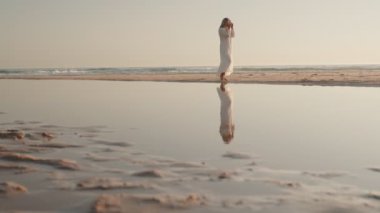 Genç, kaygısız, beyaz elbiseli bir kadın gün doğumunda sahilde yürüyor. Onun yansıması dalgaların bıraktığı suda görülebilir. Huzurlu ve güzel bir manzara..