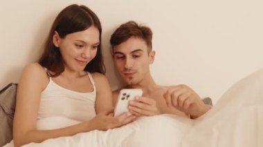 Sevecen genç çift, yatakta battaniyenin altında yatarken akıllı telefondan video seyretmeye dalmış, samimi bir anı paylaşıyorlar..