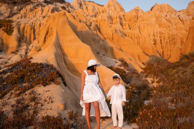 Beyaz giyinmiş bir anne ve oğul gün batımında eşsiz kaya oluşumları ve ılık renkleri ile çarpıcı bir çöl manzarasını keşfediyorlar..
