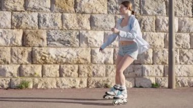 Güneşli bir günde paten kaymaktan hoşlanan genç bir kadının yan görüntüsü, sıradan bir yaz kıyafeti giymiş, taş bir duvarın yanındaki kaldırım yolunda..