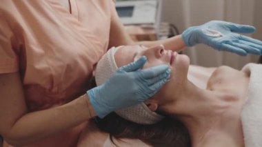 Yetenekli bir güzellik uzmanı, profesyonel bir güzellik salonundaki rahat bir kadının yüzüne nazikçe yüz masajı yapar..