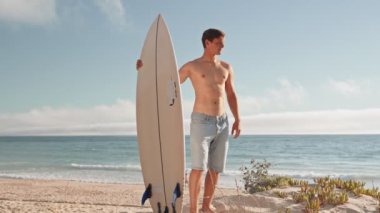 Genç bir adam güneşli bir sahilde elinde sörf tahtasıyla dalgaları seyrediyor. Rahatlamış ve kendinden emin bir ifadeyle zinde ve bronz tenli..