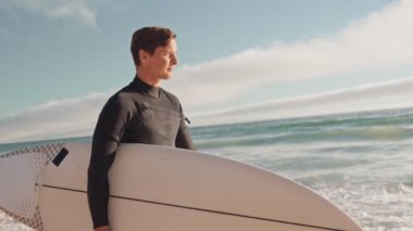 Güneşli bir günde sörf tahtasıyla sahilde yürüyen bir sörfçü. Dalgıç kıyafeti giyiyor ve sörf tahtasını kolunun altında taşıyor..