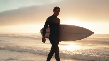 Dalgıç kıyafetli bir sörfçü gün batımında sörf tahtasıyla sahilde yürür. Batan güneşin gölgesinde kaldı..