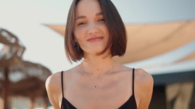 Kaygısız genç kadın saçlarında rüzgarla kameraya gülümsüyor. Kadın bir plaj otelinde..