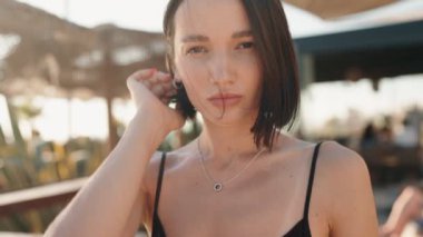 Kısa siyah saçlı, siyah bluzlu, kendine güvenen genç bir kadın saçını düzeltiyor ve kameraya bakıyor. Gün boyunca açık bir mekanda ve hafifçe gülümsüyor..