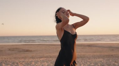 Kısa siyah saçlı kaygısız genç kadın, siyah bir elbise giyiyor. Gün batımında huzurlu bir kumsalda dinleniyor..