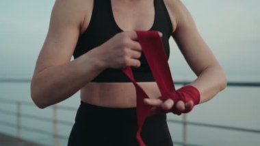 Kendini işine adamış bir kadın dışarıda boks antrenmanına hazırlanıyor. Ellerini kırmızı boks örtüsüyle kaplıyor. Kararlılığı ve gücü simgeliyor..