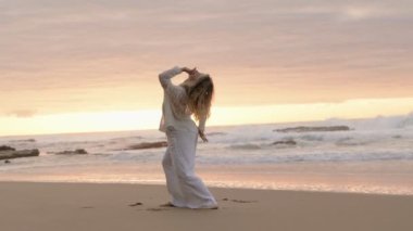 Zarif bir dansçı, günbatımında sakin bir plajda beyazlar içinde çağdaş bir dans gösterisi sergiliyor..