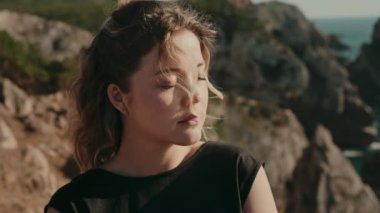 Kapalı gözlü genç bir kadın rüzgarlı bir günde arka planda dağlar ve yeşilliklerle dışarıda eğleniyor..