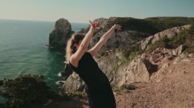 Dar siyah elbiseler içinde zarif bir şekilde çağdaş dans eden bir kadın. Resimli bir sahil manzarası arka planına karşı..