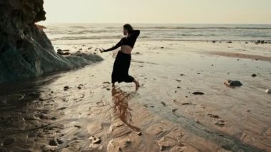 Yalnız bir dansçı, gün batımında, okyanusun yumuşak sesleri ve doğanın güzelliği ile çevrili sakin bir sahilde zarifçe yürür..