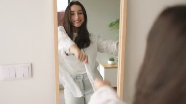 Neşeli, genç esmer kadın, günlük kıyafetlerle dans ediyor ve aynadaki yansımasına bakıp gülümsüyor..