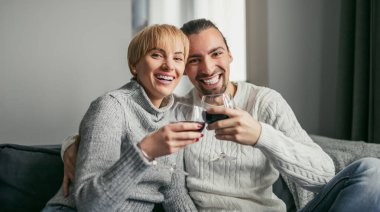 Smiling couple sitting on a couch, enjoying glasses of wine together in a cozy home setting. clipart