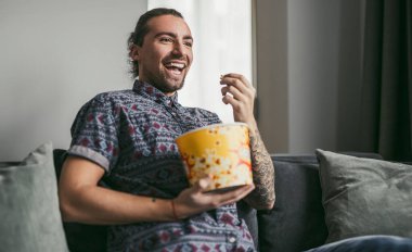 Adam evde yoğun bir filmden zevk alıyor, kanepede rahat bir şekilde otururken gülüyor ve patlamış mısır yiyor..