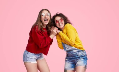 İki neşeli LGBT kadını gülüyor ve el ele tutuşuyor, günlük kıyafetler ve güneş gözlüğü takıyor, pembe arka planda duruyorlar..