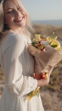 Kumsalda güneşli bir günde, beyaz elbiseli, gülümseyen ve elinde güzel bir buket çiçek tutan mutlu bir kadın. Aşk, mutluluk ve yaz temaları için mükemmel..