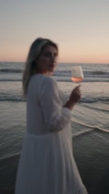 Beyaz elbiseli genç bir kadın, arkasında güneş batarken kumsalda bir bardak gül şarabı içiyor. Yaz havası, tatil, tasasız.