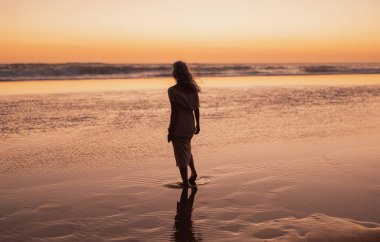 Yalnız bir kadın gün batımında kumlu sahilde yürür, titreşen gökyüzüne karşı sakin bir siluet yaratır. Huzurlu dalgalar ve altın ışık bir huzur ve yansıma hissi uyandırıyor..