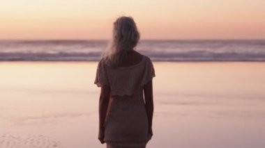 Beyaz elbiseli yaşlı bir kadın gün batımında okyanus dalgalarına doğru yürür. Emeklilik, nostalji, barış.