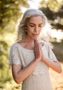 Derin meditasyonda gözleri kapalı zarif bir kadın, doğa ile çevrili. Huzurlu sahne huzuru ve farkındalığı resmediyor, doğal ortamdaki huzurlu bir anı yakalıyor..