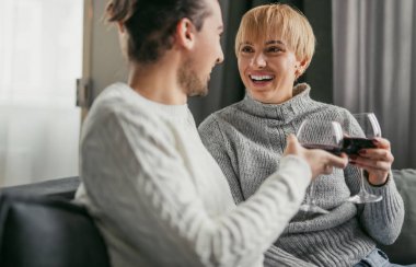 Evdeki kanepede dinlenirken kırmızı şarapla kadeh kaldıran rahat kazaklı, gülümseyen bir çift. Mutluluk ve birliktelik kavramı.