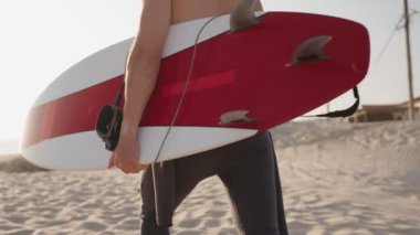 Bir sörfçü sörf tahtasıyla sahilde yürüyor, dalgaları yakalamaya hazır. Güneş batıyor, sıcak, altın bir parıltı yaratıyor..