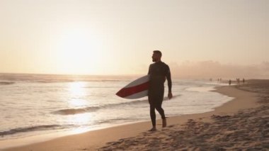Gün batımında bir sörfçü sörf tahtasını taşıyarak sahil boyunca yürür. Huzurlu atmosferin ve altın ışığın tadını çıkarıyor..