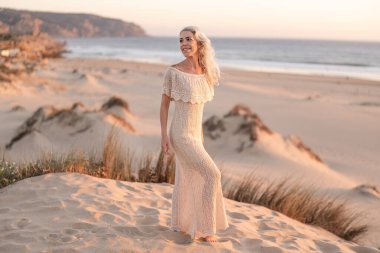 Kumlu kum tepelerinde bej bir elbise giymiş gün batımında sahile bakan bir kadın. Yumuşak parıltı, kıyı manzarasının huzur ve doğal güzelliğini vurguluyor..