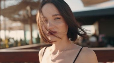 Güzel, genç, esmer bir kadın açık hava restoranında saçları rüzgarda uçuşurken kameraya gülümsüyor. O mutlu ve tatilinin tadını çıkarıyor..