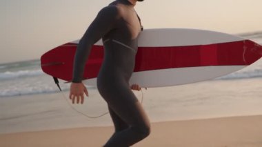 Dalgıç kıyafetli bir sörfçü gün batımında sahilde sörf tahtasını taşıyarak yürür. Akşam sörf yapmak için okyanusa girmeye hazırlanıyor..