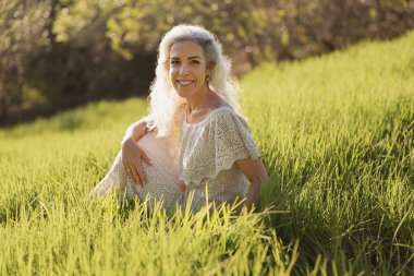 Güneşli bir çayırda dinlenen neşeli bir kadının huzurlu bir portresi. Yeşiller yemyeşil çimenler ve yumuşak ışık huzurlu ve canlandırıcı bir atmosfer yaratıyor. Rahatlık anları yakalamak için mükemmel.